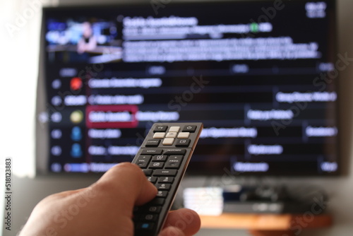 Hand with a remote control, in the background is a TV with a list of channels.