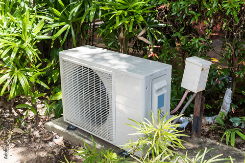 A split type inverter air conditioning condenser mounted outside a house partially concealed with shrubs. HVAC for tropical countries. photo