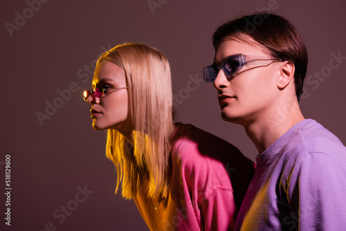 Stylish couple in sunglasses looking away isolated on purple with lighting.