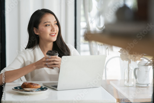 Asian woman using laptop in coffee shop and looking outside dreaming about something