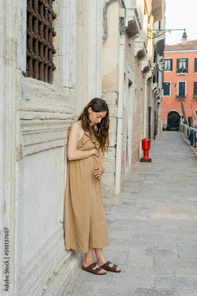 Young pregnant woman in dress touching belly on urban street in Italy.