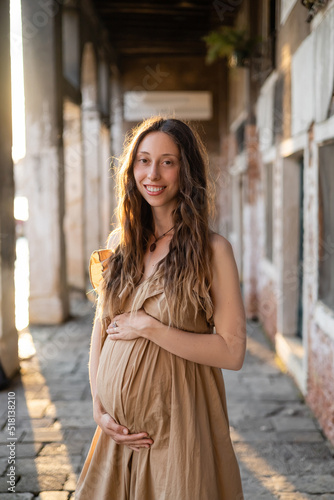 Positive pregnant woman in dress looking at camera near blurred old building in Italy.