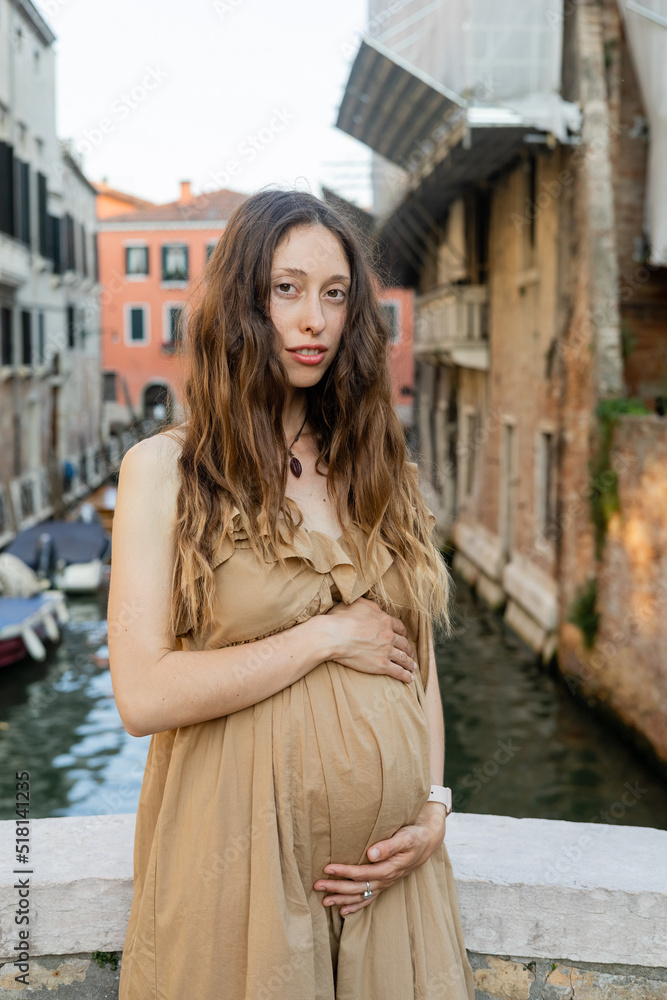Portrait of pregnant woman in dress looking at camera in blurred Venice.