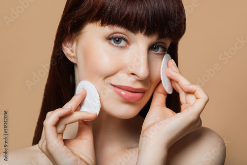 Portrait of young caucasian woman holding white cosmetic sponges and smiling on camera over beige background. Natural beauty and skin care concept. photo