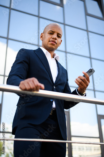 Hairless businessman holding smartphone and looking at camera near building on street 