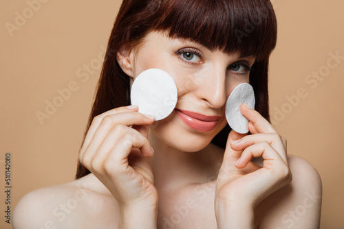 Portrait of young caucasian woman holding white cosmetic sponges and smiling on camera over beige background. Natural beauty and skin care concept. photo