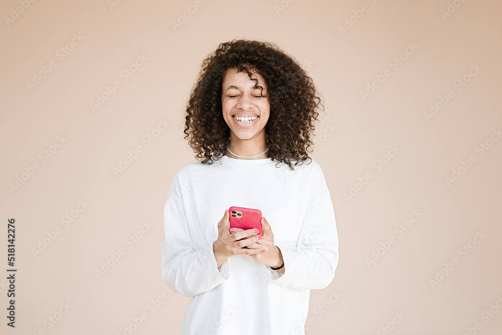 Adorable african american young woman holds mobile phone, browses social networks, has broad smile, enjoys online chatting, isolated on beige background, being always in touch Adorable african american young woman holds mobile phone, browses social networks, has broad smile, enjoys online chatting, isolated on beige background, being always in touch