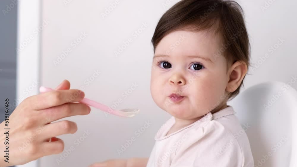 Baby girl eating blend mashed food sitting, on high chair, mother feeding child, hand with spoon for vegetable lunch, baby weaning, first solid food for young kid. Baby girl eating blend mashed food sitting, on high chair, mother feeding child, hand with spoon for vegetable lunch, baby weaning, first solid food for young kid.