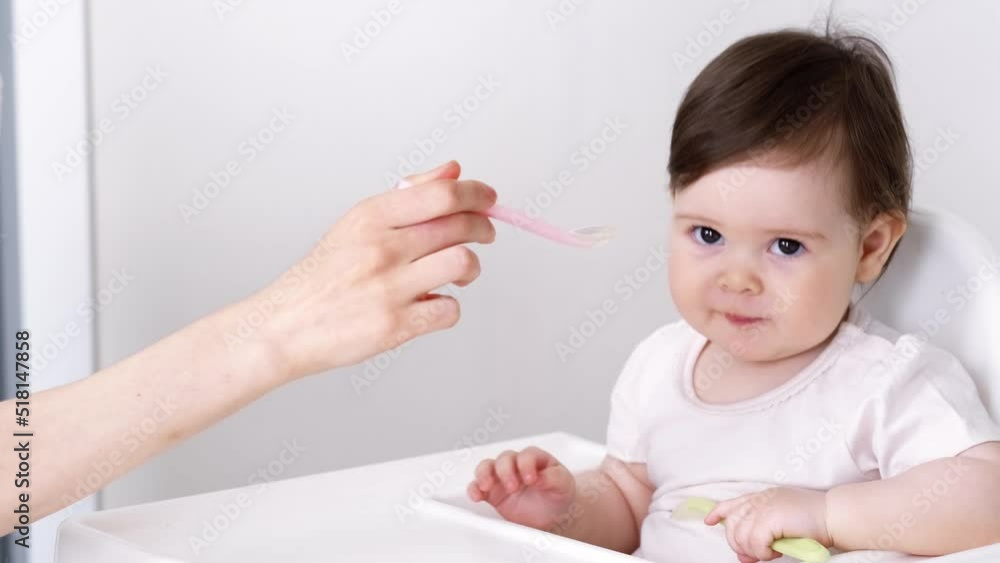 Baby girl eating blend mashed food sitting, on high chair, mother feeding child, hand with spoon for vegetable lunch, baby weaning, first solid food for young kid. Baby girl eating blend mashed food sitting, on high chair, mother feeding child, hand with spoon for vegetable lunch, baby weaning, first solid food for young kid.