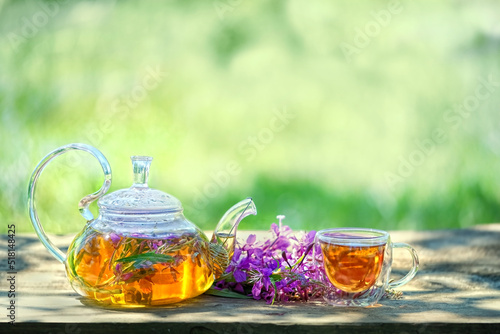 Glass teapot and cup with tea from flowers of fireweed on wooden table in garden, abstract green natural background. Useful herbal tea, Traditional Koporye Tea Decoction, infusion of kiprey, ivan tea.