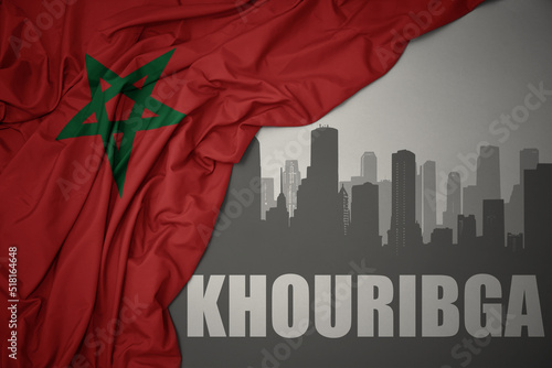 abstract silhouette of the city with text Khouribga near waving colorful national flag of morocco on a gray background.