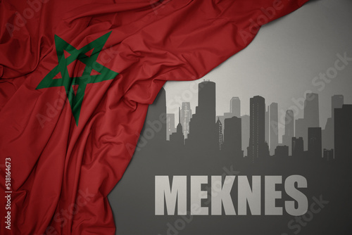 abstract silhouette of the city with text Meknes near waving colorful national flag of morocco on a gray background.