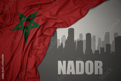 abstract silhouette of the city with text Nador near waving colorful national flag of morocco on a gray background. photo