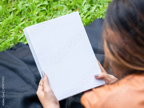 Mockup of a white blank hardcover book featuring a woman reading it at a park