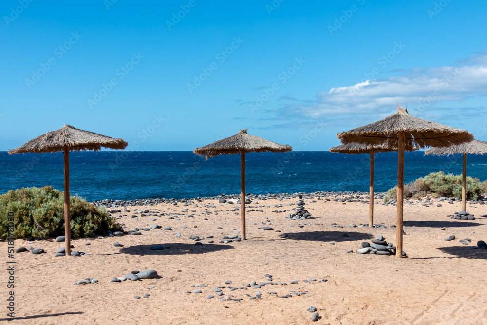 Straw sun umbrellas on sand beach connected to stone pebble beach Playa de San Blas near coastal village Los Abrigos, Tenerife, Canary Islands, Spain, Europe, EU. Coastline of Atlantic Ocean. Vacay