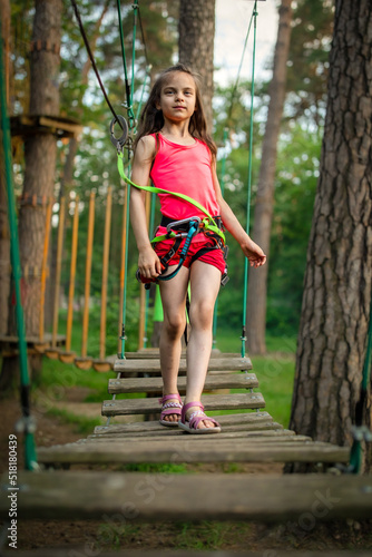 Little smiling girl in harness passing on trail for kids in sky rope park in summer