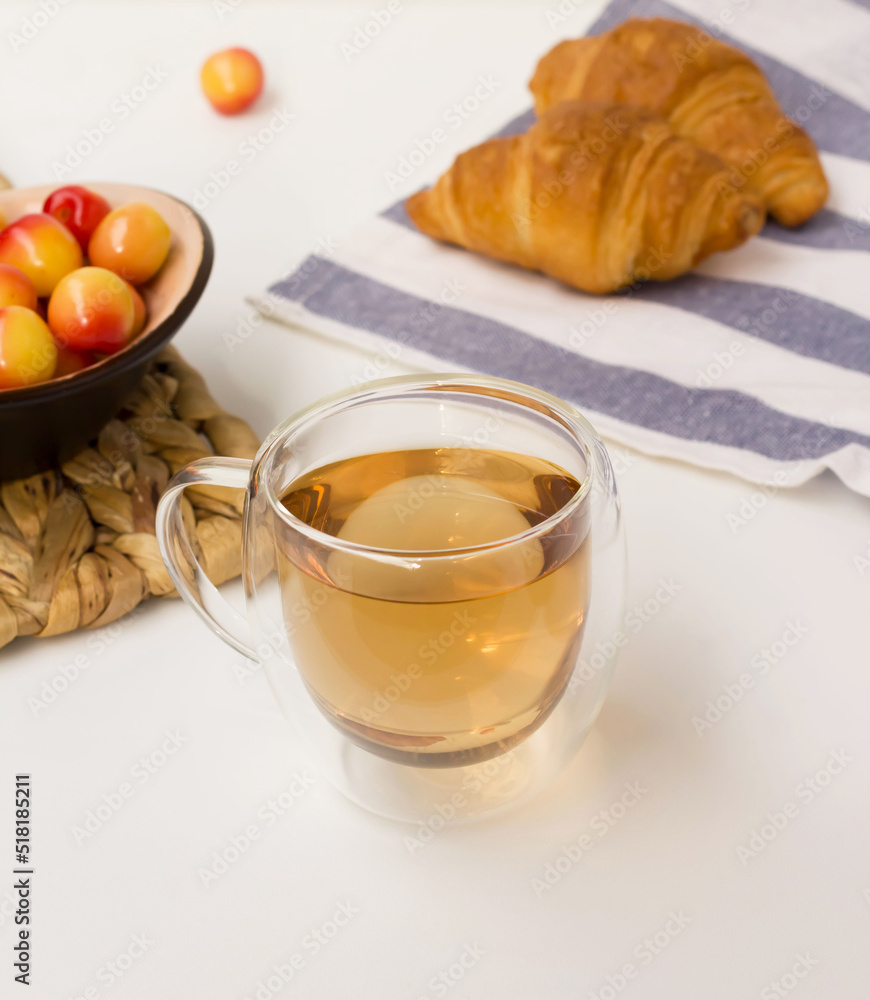 Herbal tea in a glass cup on a white surface. Yellow-red cherries and two croissants on a linen towel Herbal tea in a glass cup on a white surface. Yellow-red cherries and two croissants on a linen towel