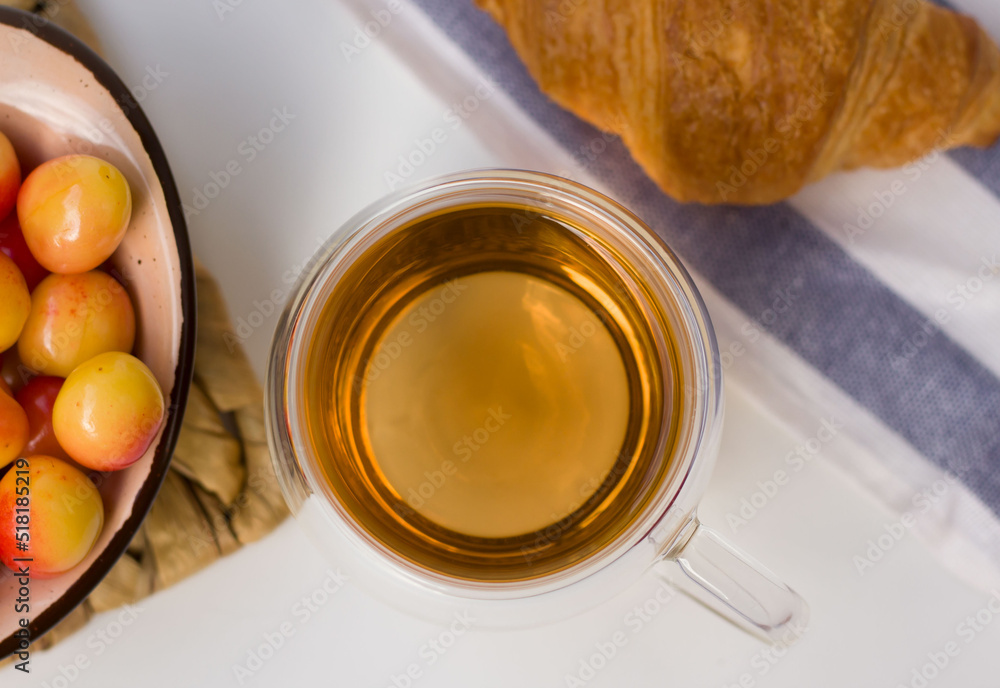 Top view of herbal tea in glass cup on white surface. Summer breakfast with cherries and croissants Top view of herbal tea in glass cup on white surface. Summer breakfast with cherries and croissants