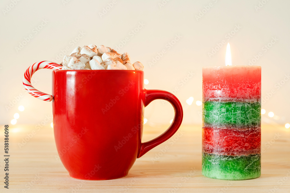 Christmas candle and cocoa in red mug with marshmallows and candy cane on wooden table with holiday lights. Hot chocolate, cacao in cup, winter drink. Christmas candle and cocoa in red mug with marshmallows and candy cane on wooden table with holiday lights. Hot chocolate, cacao in cup, winter drink.