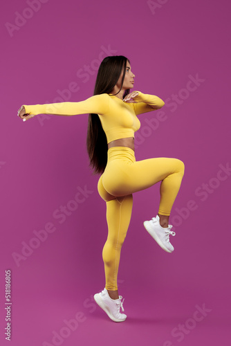 Full lengh image young fit brunette woman in yellow sport outfit jumping over purple background.