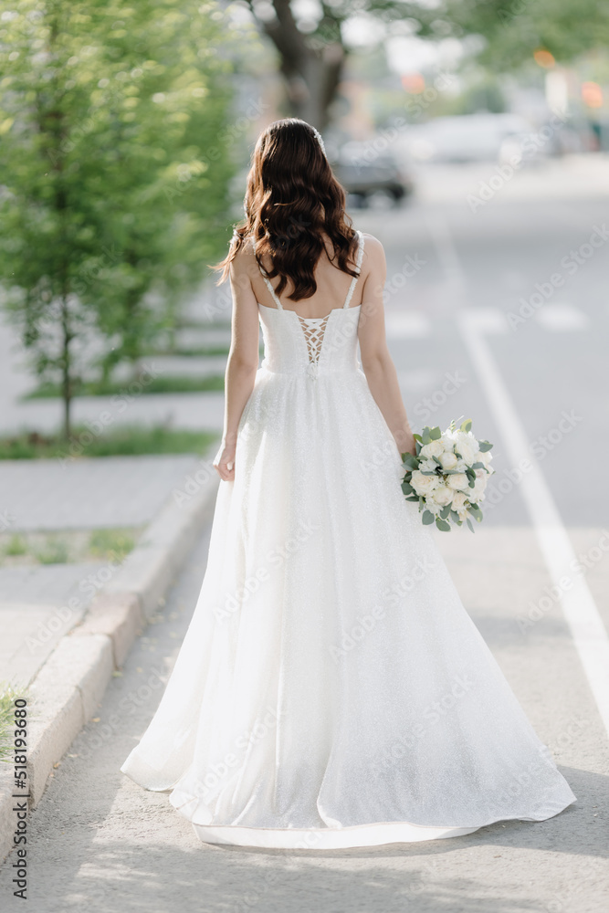 A stylish European bride in a white dress with a white bouquet is standing on the street. Photo from the back