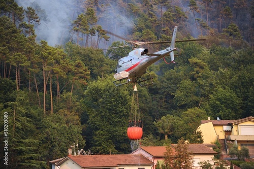 Un elicottero sta portando un bucket con acqua per spengere il fuoco tra il bosco e le case, Toscana, Italia