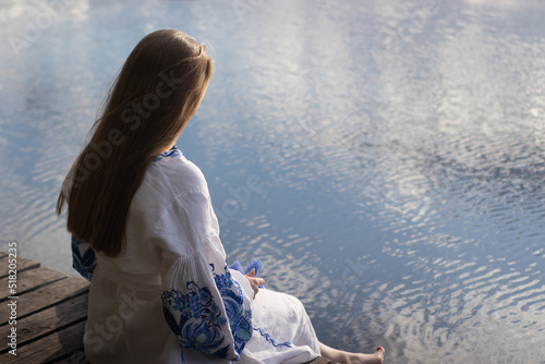 a girl in an embroidered Ukrainian shirt sits on the pier, the reflection of clouds in the water of the lake. On the shore of the sky. vyshyvanka day. freedom. patriot photo