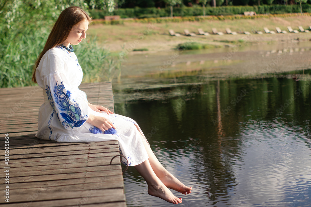 a girl in an embroidered Ukrainian shirt sits on the pier, the reflection of clouds in the water of the lake. On the shore of the sky. vyshyvanka day. freedom. patriot a girl in an embroidered Ukrainian shirt sits on the pier, the reflection of clouds in the water of the lake. On the shore of the sky. vyshyvanka day. freedom. patriot