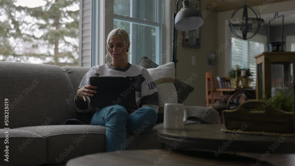 Woman sitting on the couch in front of a bay window, drinking coffee while scrolling on a tablet sitting straight up. Filmed from the front side. Woman sitting on the couch in front of a bay window, drinking coffee while scrolling on a tablet sitting straight up. Filmed from the front side.