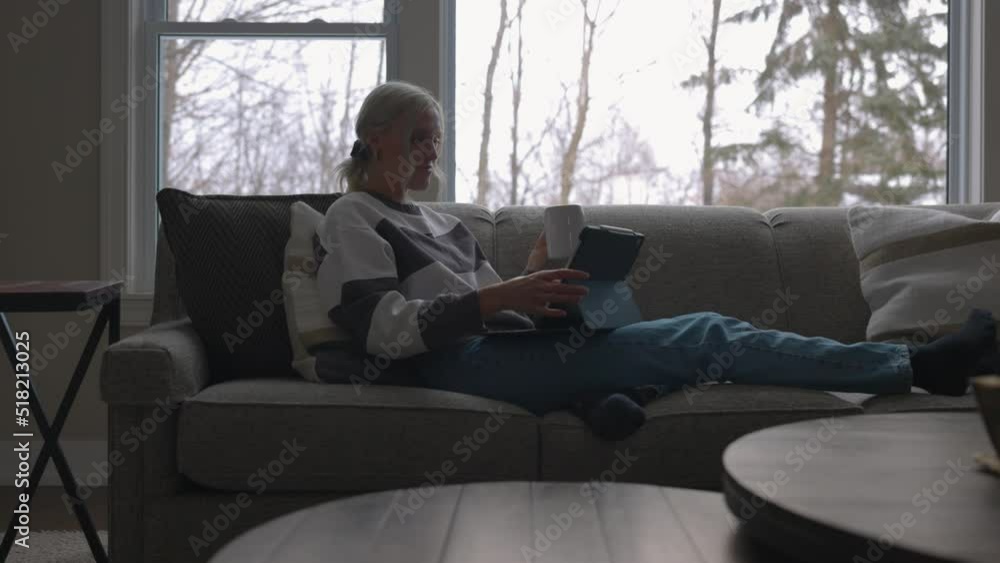 Woman sitting on the couch in front of a bay window, drinking coffee while scrolling on a tablet close up from the front. Woman sitting on the couch in front of a bay window, drinking coffee while scrolling on a tablet close up from the front.