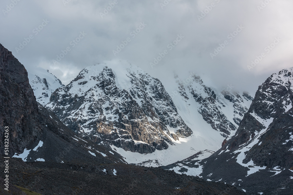 Dramatic landscape with sunlit snowy mountain range in low cloudy sky. Golden shine on white snow and black rocks in low clouds. Awesome view to snow mountains with gold sunlight in changeable weather