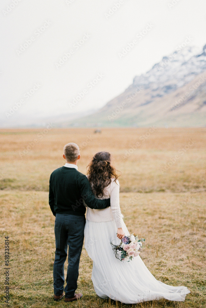 Groom hugs bride with a bouquet on a dry meadow at the foot of the mountains. Back view