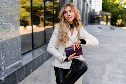 Sucsessful blond woman with perfect wavy hairs in casual outfit with luxury purse posing over modern business center.