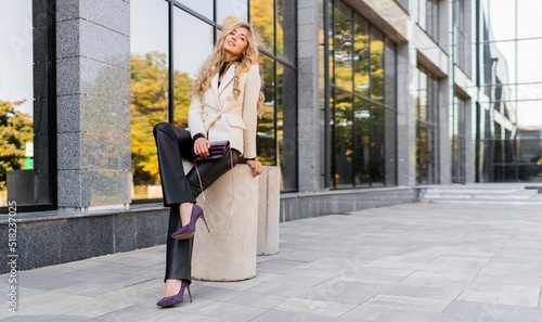 Outdoor photo of sucsessful blond woman with perfect wavy hairs in casual outfit with luxury purse posing over modern business center.