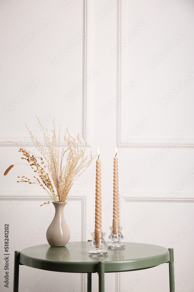 Dry plants in vase and burning candles on table near white wall