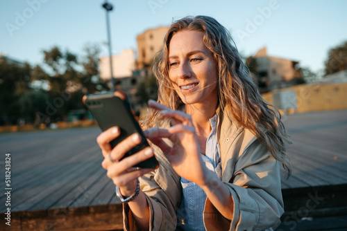 Young casual woman in coat sitting on bench on sea embankment using smartphone