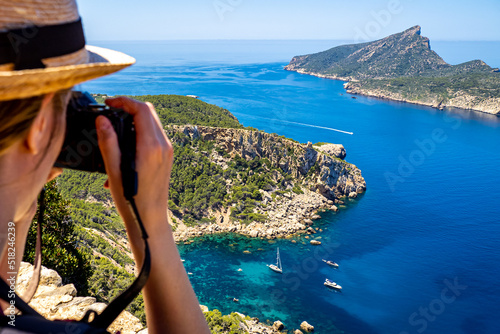 Fotobehang A female photographer with hat shots a picture from a cliff of the rocky coastline of idyllic Mallorca cove Cala en basset nearby Sant Elm with boats and the island La Dragonera in the background