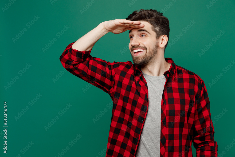 Young happy smiling caucasian man he 20s wearing red shirt grey t-shirt hold hand at forehead look far away distance isolated on plain dark green background studio portrait People lifestyle concept.