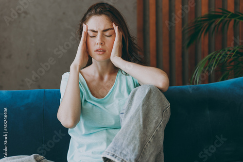 Young sad sick ill caucasian woman she 20s wearing casual clothes mint t-shirt holding head rub temples suffer from headache make solution sit on blue sofa indoor rest at home in own room apartment photo