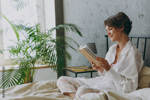 Side view young calm happy woman wear white shirt pajama she lying in bed read book study rest relax spend time in bedroom lounge home in own room hotel wake up dream be lost in reverie good mood day photo