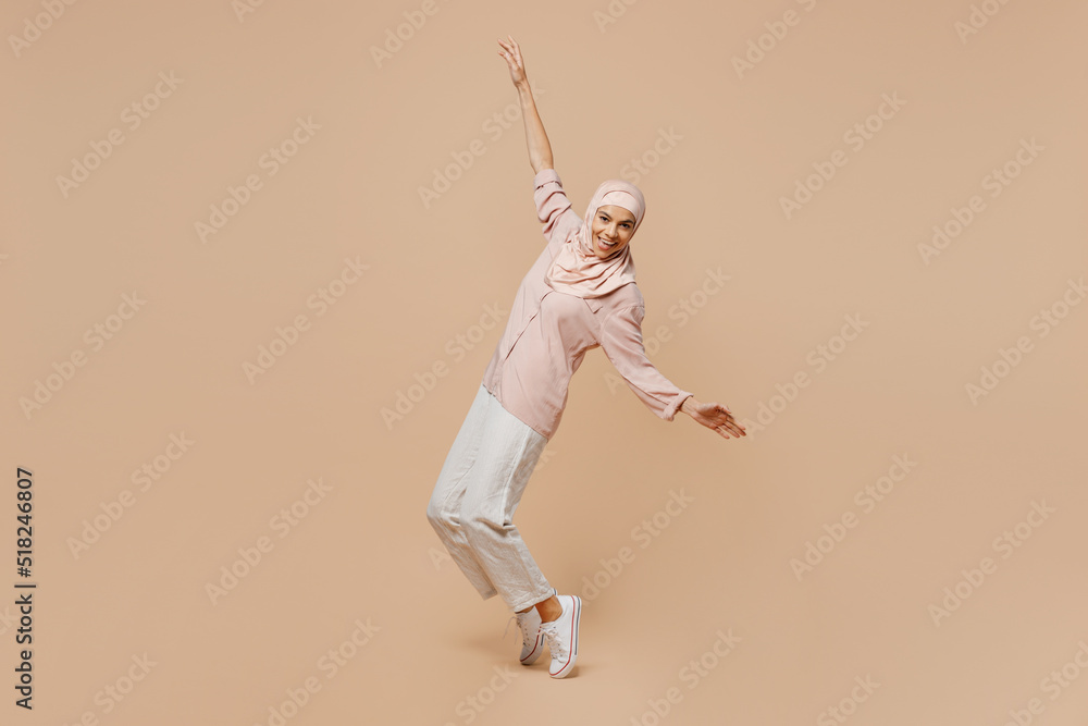 Full body young arabian asian muslim woman she wear abaya hijab pink clothes stand on toes with outstretched hands leaning back dance isolated on plain beige background People islam religious concept