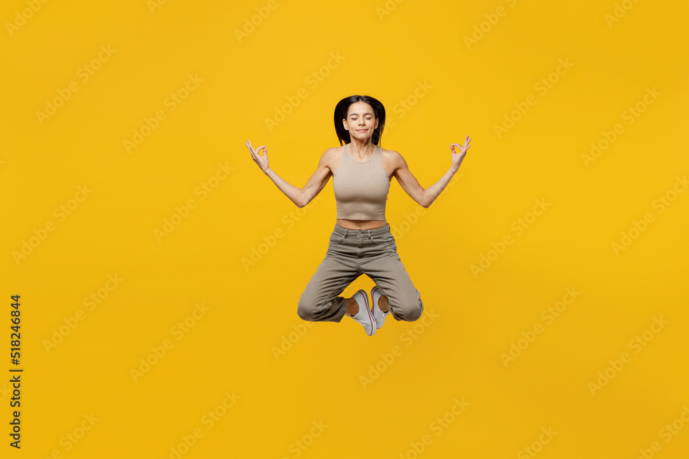 Full body young tranquil spiritual latin woman 30s she wear beige tank shirt jump high hold spread hands in yoga om aum gesture relax meditate try calm down isolated on plain yellow backround studio.