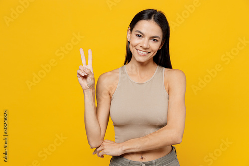 Young smiling friendly positive fun cool cheerful latin woman 30s she wearing basic beige tank shirt showing victory sign isolated on plain yellow backround studio portrait. People lifestyle concept.