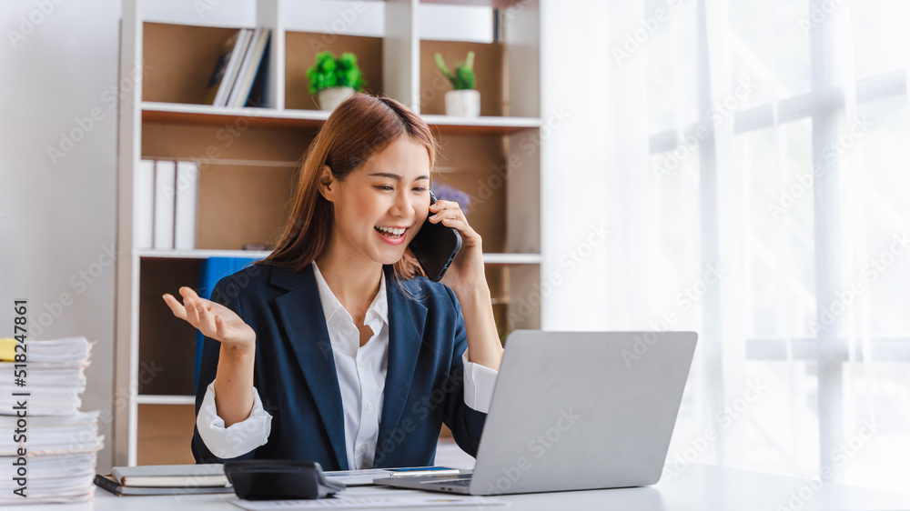 Young Attractive Asian business woman talking on mobile phone and working on laptop in modern office with financial reports. Young Attractive Asian business woman talking on mobile phone and working on laptop in modern office with financial reports.
