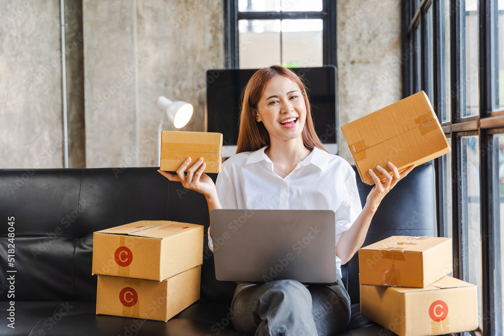Fototapeta premium Young Asian business woman on sofa using laptop computer checking customer order online shipping boxes at home.
