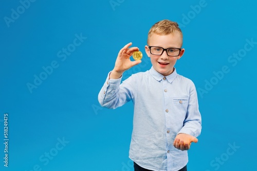 Cryptocurrency and modern finance. Cheerful boy in eyeglasses holding golden bitcoin in studio on blue background.