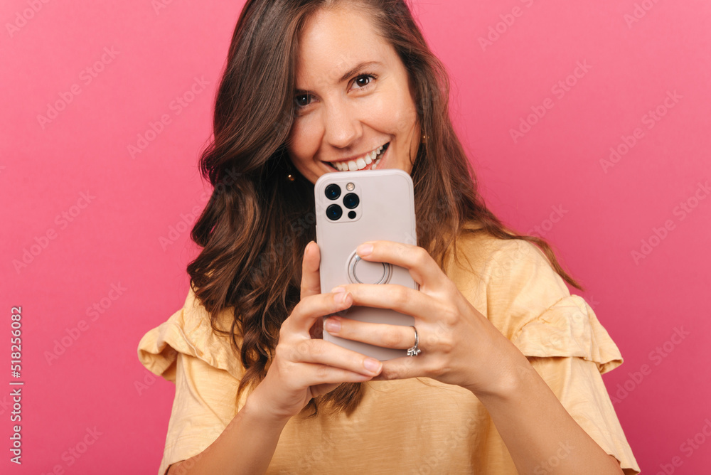 A young cheerful woman taking a photo of you with her phone is smiling sitting near a pink wall