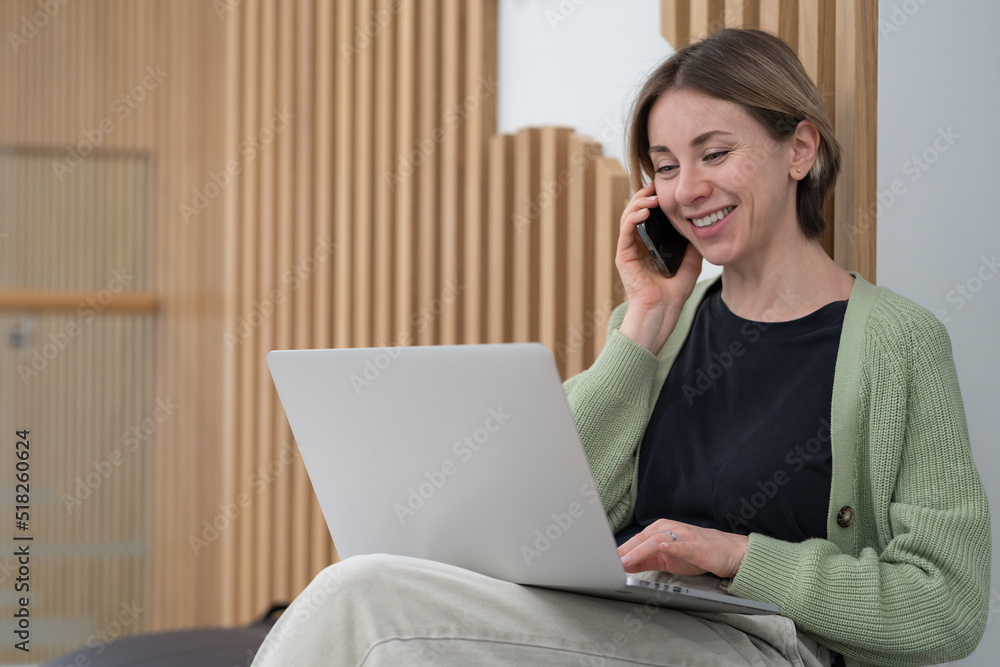 Pleased happy middle-aged businesswoman using mobile device for work communication, looking at laptop screen and communicating with client via smartphone, solving issues remotely. Freelance concept Pleased happy middle-aged businesswoman using mobile device for work communication, looking at laptop screen and communicating with client via smartphone, solving issues remotely. Freelance concept