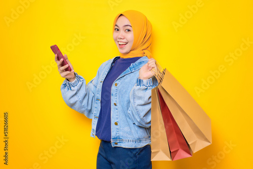 Happy young Asian woman in jeans jacket holding mobile phone and shopping bags, looking at camera isolated over yellow background