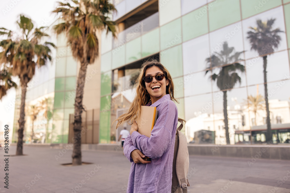 Happy young caucasian girl smiles, holds notebooks in her hands standing outdoors. Brown-haired woman wears sunglasses, shirt and backpack. City life concept